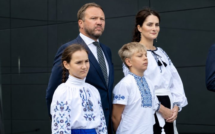 Participants of the flag-raising ceremony in Tallinn: a man in a dark suit, two children and a woman in embroidered shirts