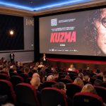 Kino Sõprus täissaal: vaatajad vaatavad suurele ekraanile filmi „Kuzma“ plakatiga, laval esineb saatejuht